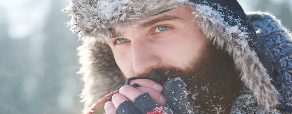 Homme barbu aux yeux clairs portant une chapka en fourrure sous la neige, réchauffant ses mains gantées en plein hiver glacial.