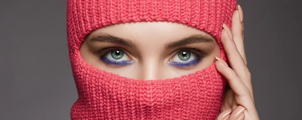 Gros plan d’une femme portant une cagoule femme rose tricotée, laissant apparaître ses yeux verts maquillés de bleu, dans un environnement neutre avec un fond gris.