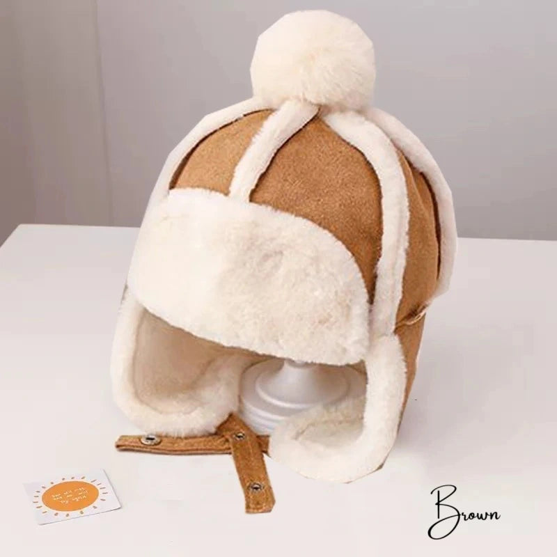 Bonnet Chapka Enfant marron avec intérieur en fausse fourrure blanche, pompon en laine et oreillettes rabattables, posé sur un support blanc dans un cadre minimaliste.