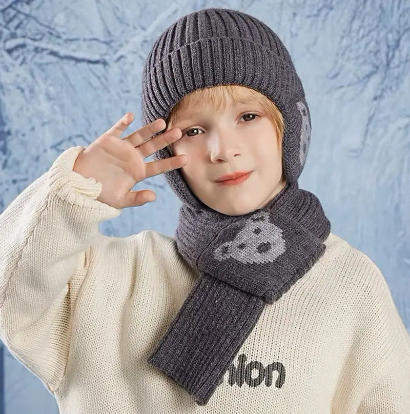 Garçon portant un ensemble bonnet écharpe enfant en tricot gris foncé avec des motifs d'ours, saluant avec la main, vêtu d'un pull beige, sur un fond givré évoquant l'hiver.