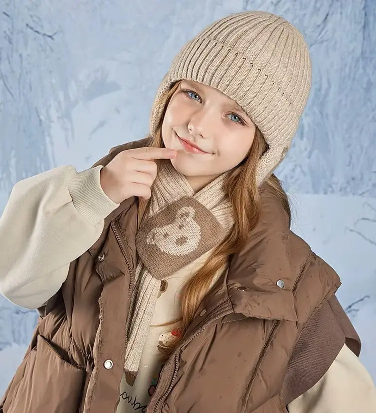 Fille portant un ensemble bonnet écharpe enfant en tricot beige avec des motifs d'ours, vêtue d'une veste matelassée marron et d'un pull clair, posant avec un sourire sur un fond hivernal givré.