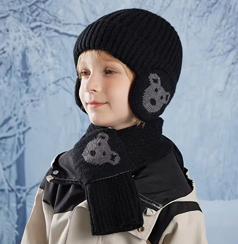 Garçon portant un ensemble bonnet écharpe enfant en tricot noir avec des motifs d'ours gris, vêtu d'une veste d'hiver noire et beige, posant devant un fond givré évoquant un paysage hivernal.