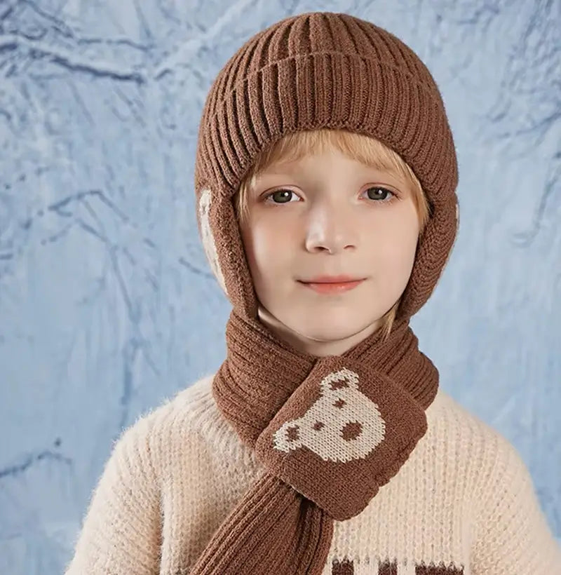 Garçon portant un ensemble bonnet écharpe enfant en tricot marron avec des motifs d'ours beige, vêtu d'un pull clair texturé, posé devant un fond bleu glacé évoquant l'hiver.