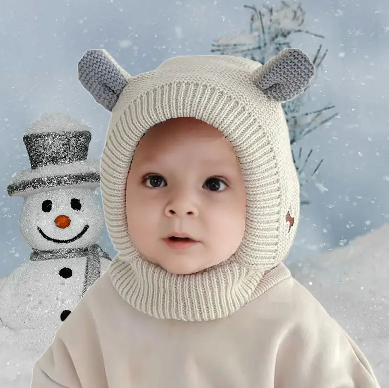 Bébé habillé chaudement avec une cagoule bébé 6 à 24 mois en tricot gris et blanc, avec de petites oreilles. Arrière-plan enneigé avec un bonhomme de neige, idéal pour l'hiver.