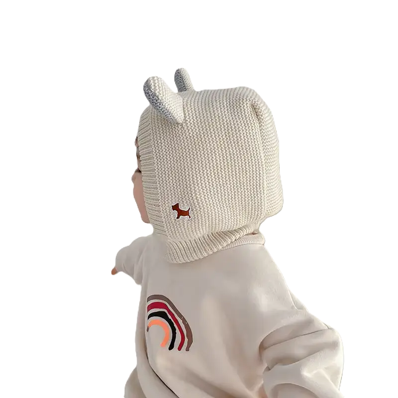 Vue latérale d'un bébé portant une cagoule bébé 6 à 24 mois blanche avec des oreilles tricotées. Détail du col montant et du motif brodé sur l’épaule.