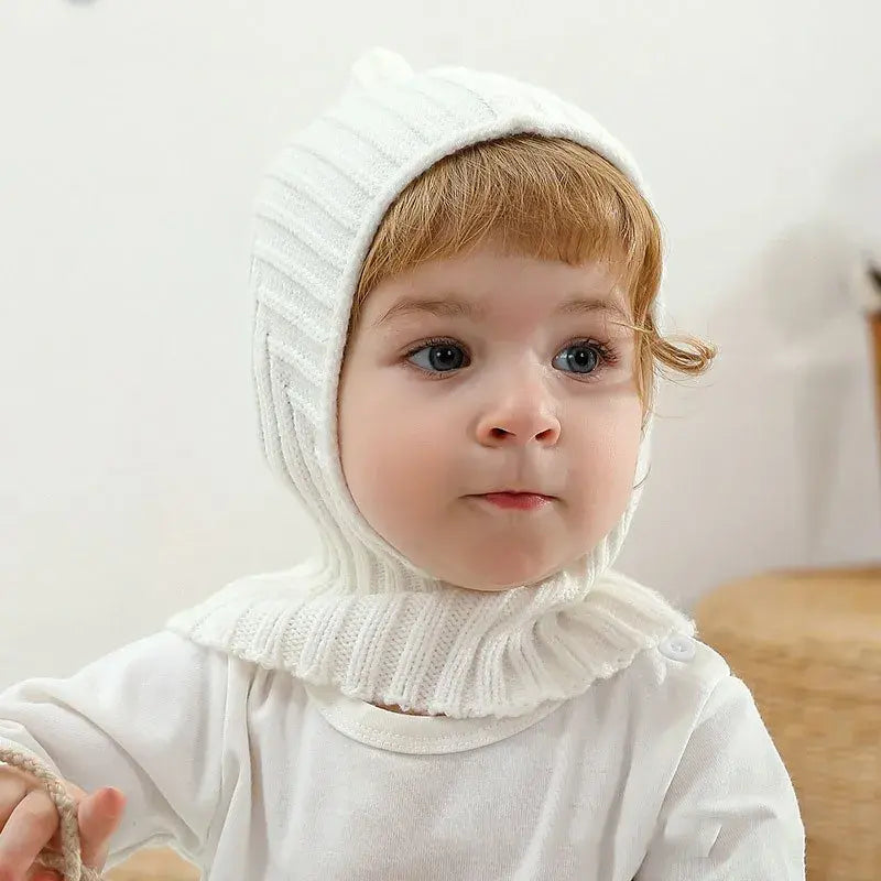 Bébé portant une cagoule blanche en tricot avec un design ajusté, couvrant la tête et le cou, parfait pour protéger du froid. Cagoule bébé adaptée aux activités hivernales.