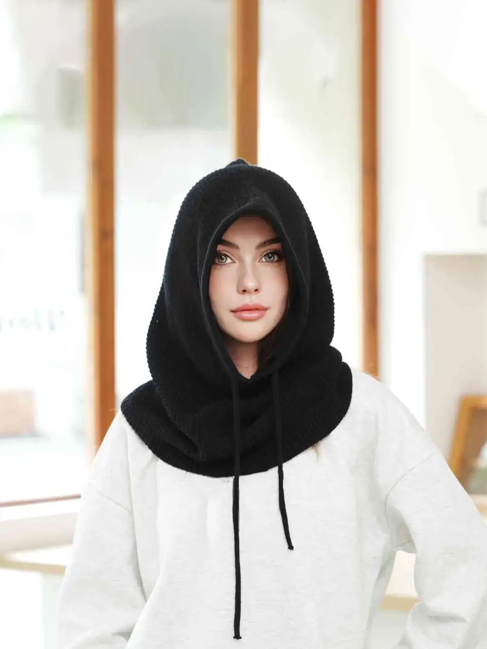 Femme portant une cagoule cache cou femme noire, capuche relevée avec cordons ajustables, offrant chaleur et protection dans un intérieur moderne.