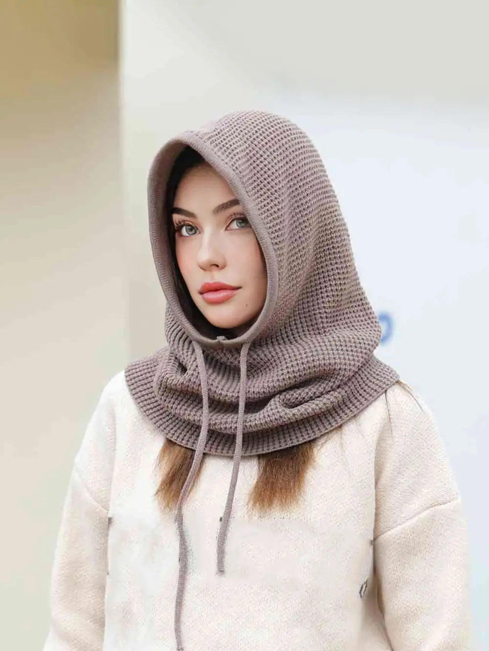 Femme portant une cagoule cache cou femme taupe, capuche relevée avec cordons ajustables, offrant élégance et protection contre le froid en extérieur.