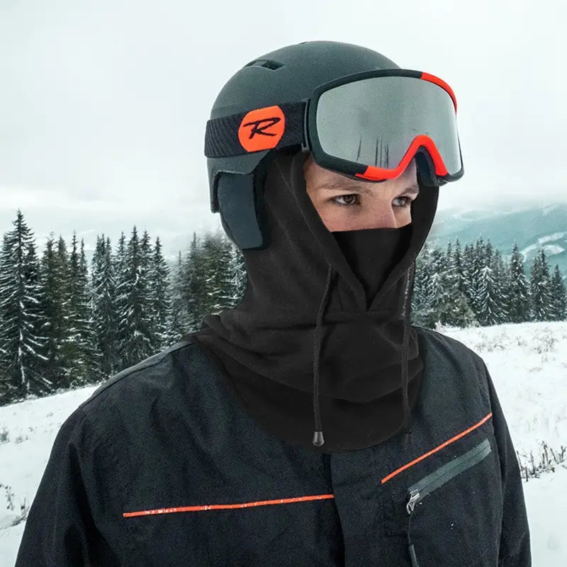 Personne équipée d'une cagoule capuche grise sous un casque de ski, avec des lunettes de protection aux reflets rouges. Le design couvrant protège le visage et le cou contre le froid extrême. Paysage enneigé en arrière-plan avec des sapins sous un ciel gris.