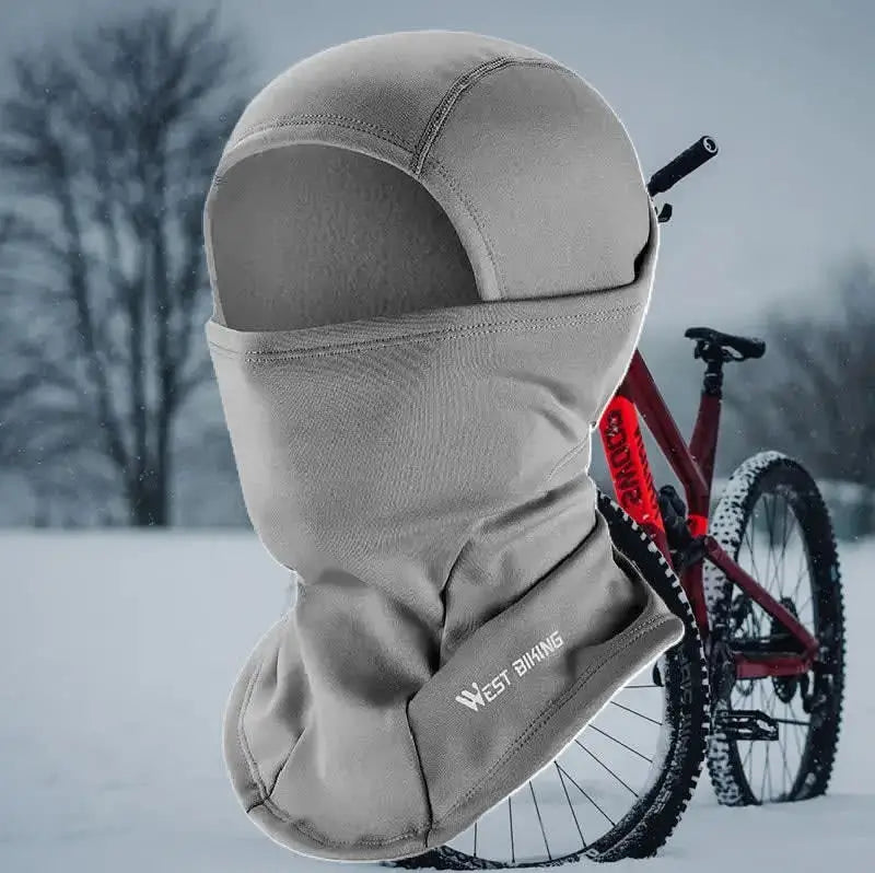 Cagoule cyclisme grise conçue pour les sorties hivernales, avec doublure chaude et ajustement ergonomique, présentée devant un vélo rouge sur un chemin enneigé.