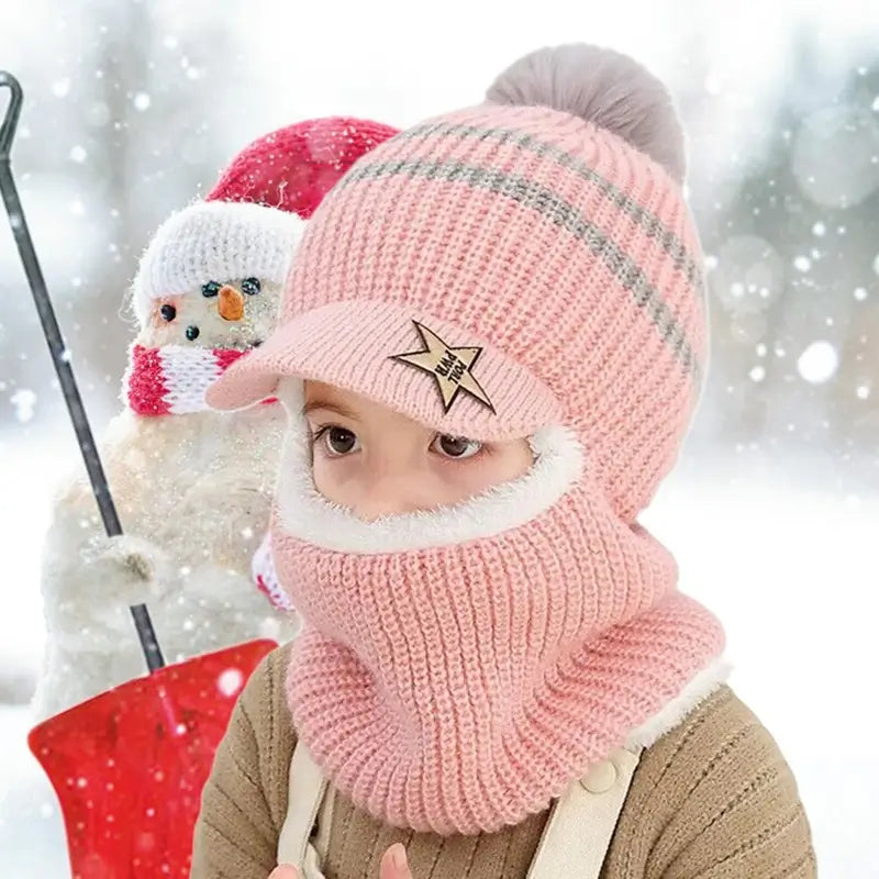 Enfant portant une cagoule enfant tricotée rose avec visière et pompon gris, couvrant le visage et le cou, devant un bonhomme de neige avec écharpe rouge, dans un paysage hivernal enneigé.