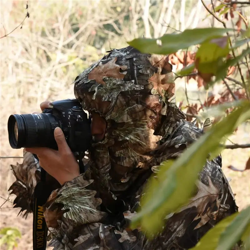 Personne portant une cagoule filet camouflage dans un environnement naturel, tenant un appareil photo reflex pour photographier la faune. La cagoule filet camouflage offre une discrétion optimale pour les activités de chasse ou d'observation en plein air.