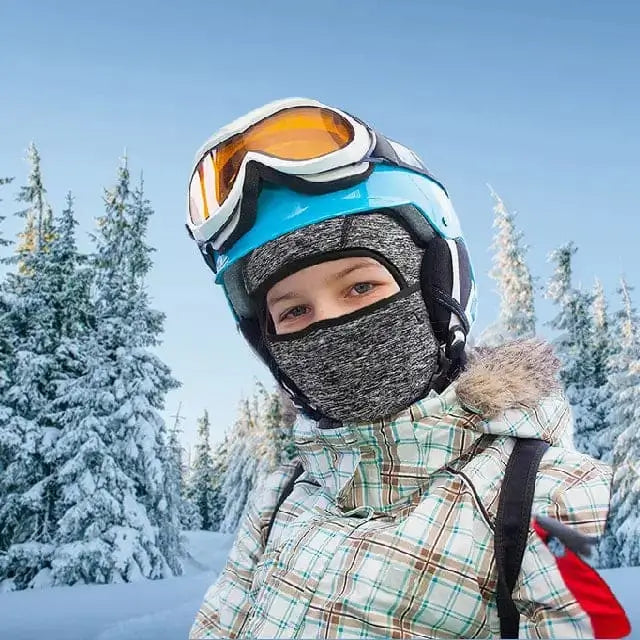 Enfant portant une cagoule fine sous casque enfant grise pendant une sortie au ski, avec des arbres enneigés et un ciel clair en arrière-plan.