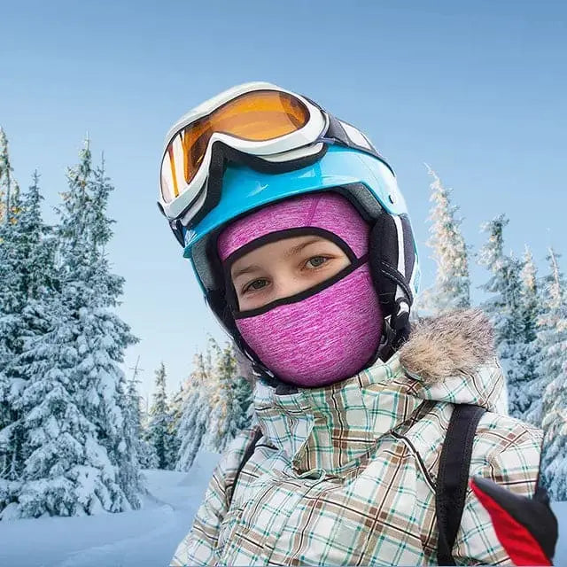 Enfant portant une cagoule fine sous casque enfant rose pendant une sortie au ski, avec des arbres enneigés en arrière-plan.