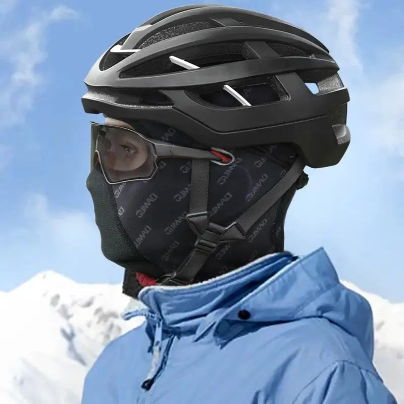 Cagoule hiver vélo noire avec motifs tribaux et zone respirante en maille, conçue pour protéger le visage et la tête des cyclistes contre le froid et le vent, présentée sur fond montagneux enneigé.