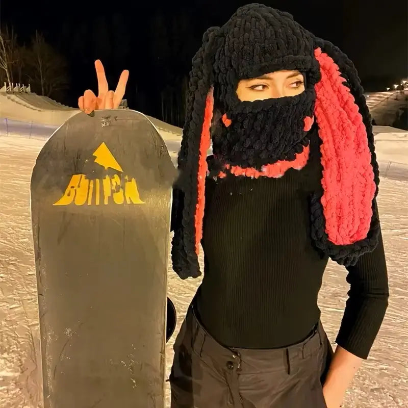 Cagoule Oreilles de lapin noire et rouge – Une snowboarder en tenue noire porte une cagoule avec des oreilles de lapin rouges. La neige est visible en arrière-plan, créant une ambiance sportive hivernale.