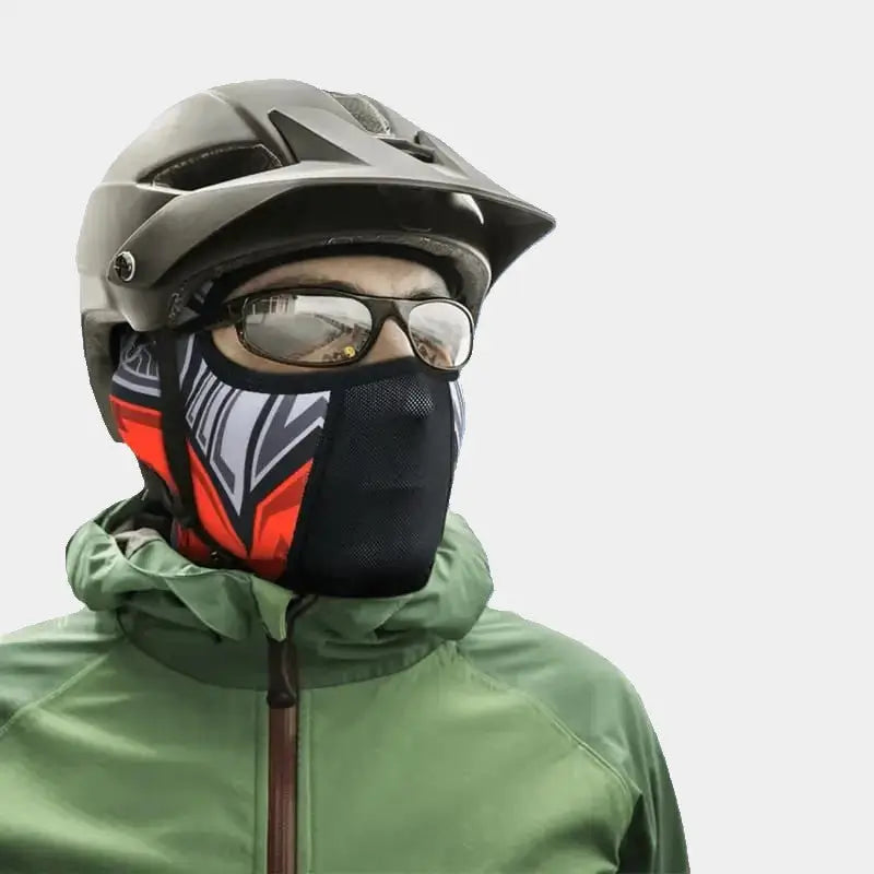 Personne portant une cagoule pour faire du vélo avec un design coloré rouge et noir, un masque respirant, et un casque gris, idéal pour les activités en plein air et les conditions froides.