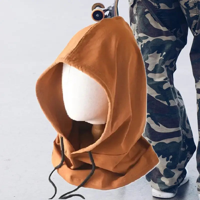 Cagoule streetwear marron avec capuche ample et cordons ajustables, posée sur un mannequin blanc. À côté, une personne en pantalon cargo camouflage et skateboard renforce l’ambiance urbaine et décontractée. Un accessoire tendance parfait pour un look streetwear authentique.