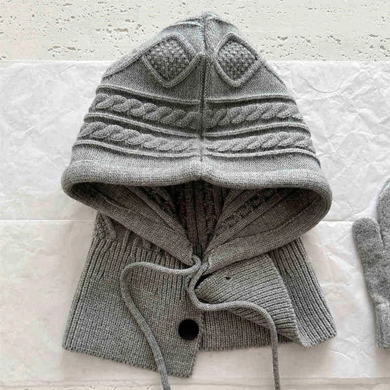 Capuche cagoule tricot grise avec motifs torsadés et cordons ajustables, posée à plat sur une surface blanche