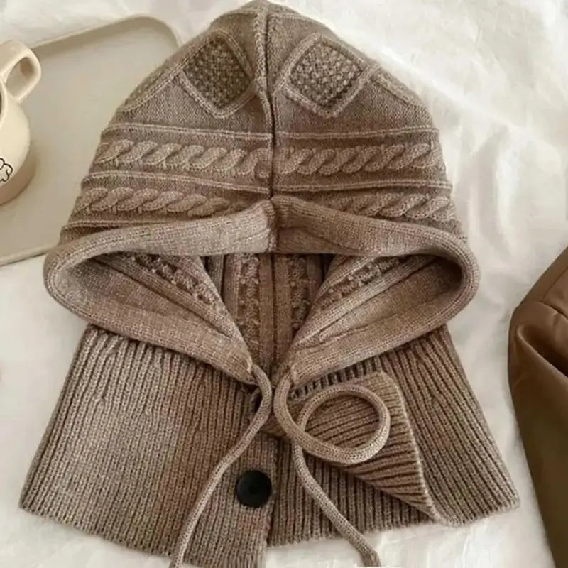 Capuche cagoule tricot kaki élégante avec détails en maille torsadée, posée à plat avec des accessoires en arrière-plan.