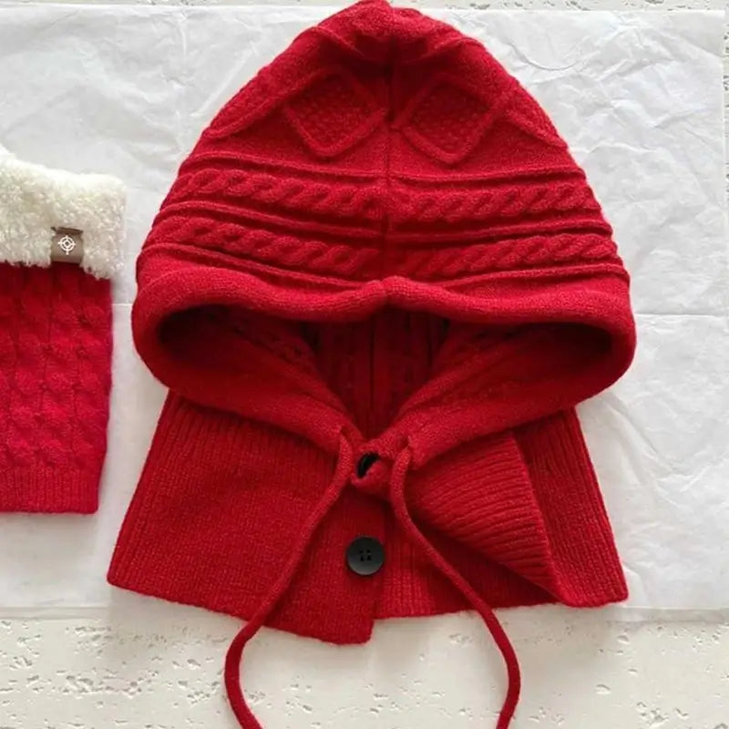 Capuche cagoule tricot rouge avec détails torsadés, cordons ajustables et bouton décoratif, posée à plat sur une surface blanche.