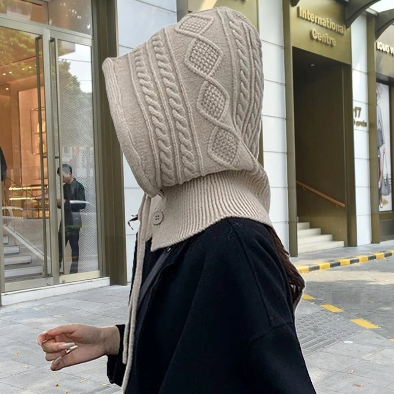 Capuche cagoule tricot beige avec motif torsadé, portée de profil par une femme dans une rue enneigée.