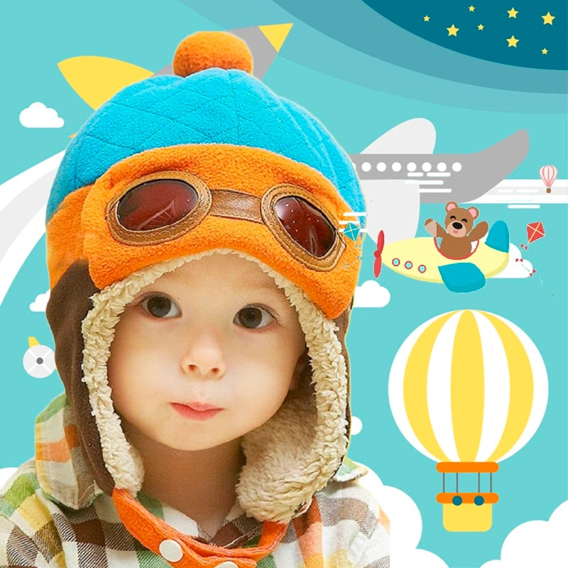 Enfant avec une chapka enfant aviateur bleue et orange, doublure chaude et lunettes, sur fond de décor aérien avec montgolfière.