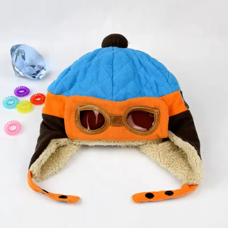 Chapka enfant aviateur bleue, orange et marron avec lunettes d'aviateur, posée sur fond blanc et décorée de boutons colorés.