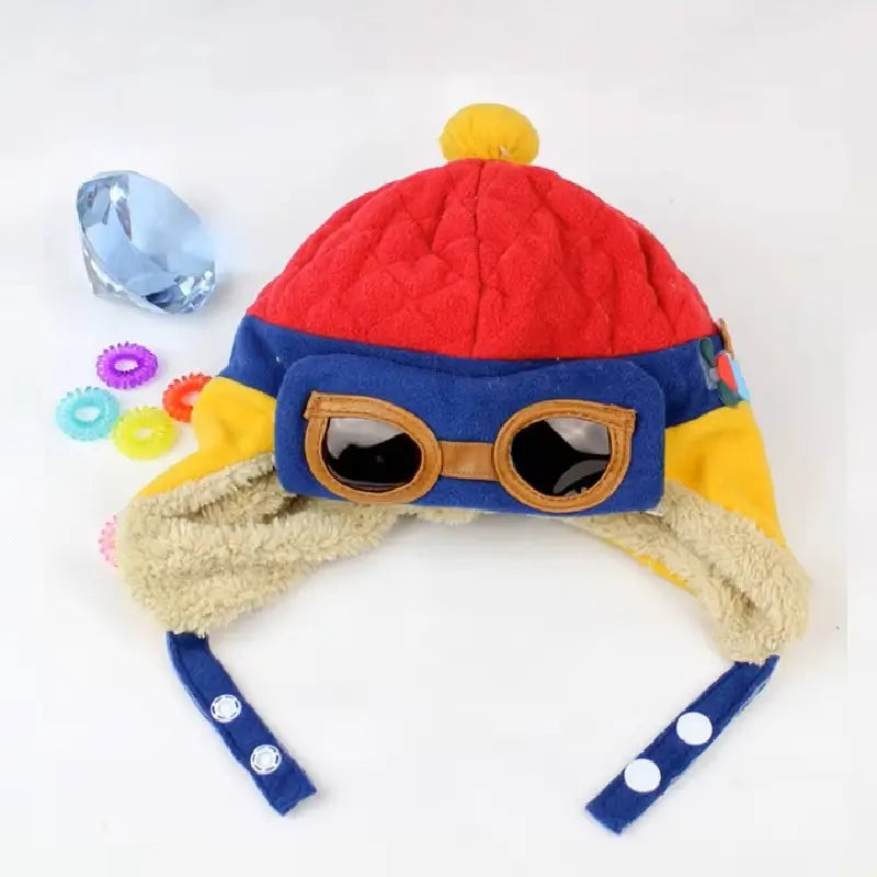 Chapka enfant aviateur bleue et rouge avec lunettes d'aviateur, pompon jaune et doublure polaire, accompagnée de perles décoratives sur fond blanc.