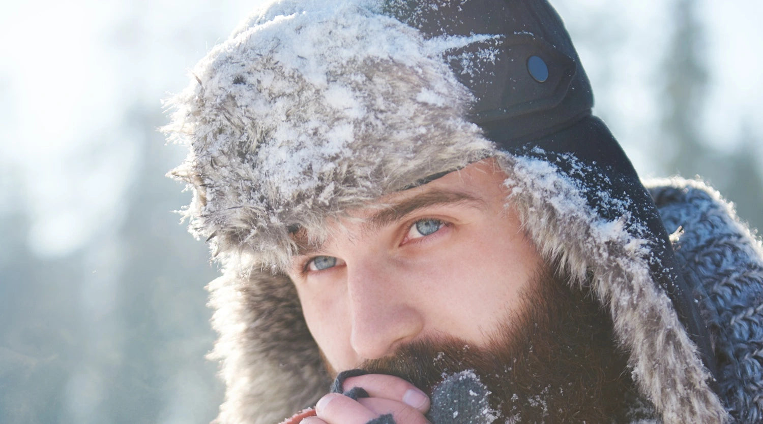 Homme barbu portant une chapka en fausse fourrure couverte de neige, illustrant la protection et la chaleur des chapkas de la collection Espace Cagoules pour affronter l'hiver.